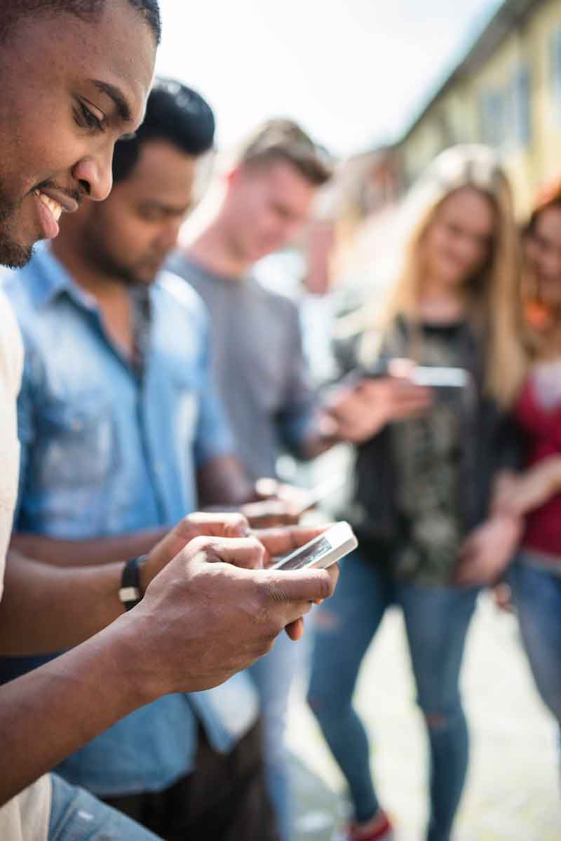 Group of five diverse people, three men, two women, all using a mobile device, with an African-American man smiling in focus in the foreground using a smartphone