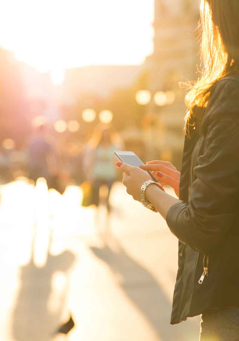 A very sunny street scene out of focus with an over-the-shoulder partial view of a woman holding an iPhone in focus on the right side of the screen